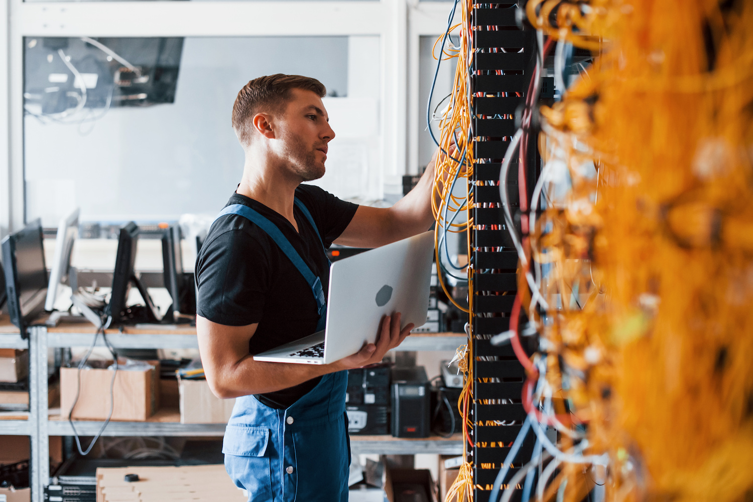 Young man in uniform and with laptop works with internet equipment and wires in server room.